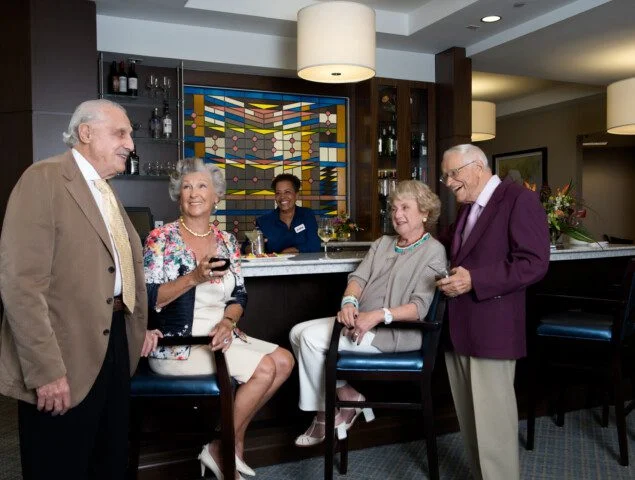 two smiling senior couples in elegant evening attire enjoy wine and conversation in front of the bar at Harbour's Edge