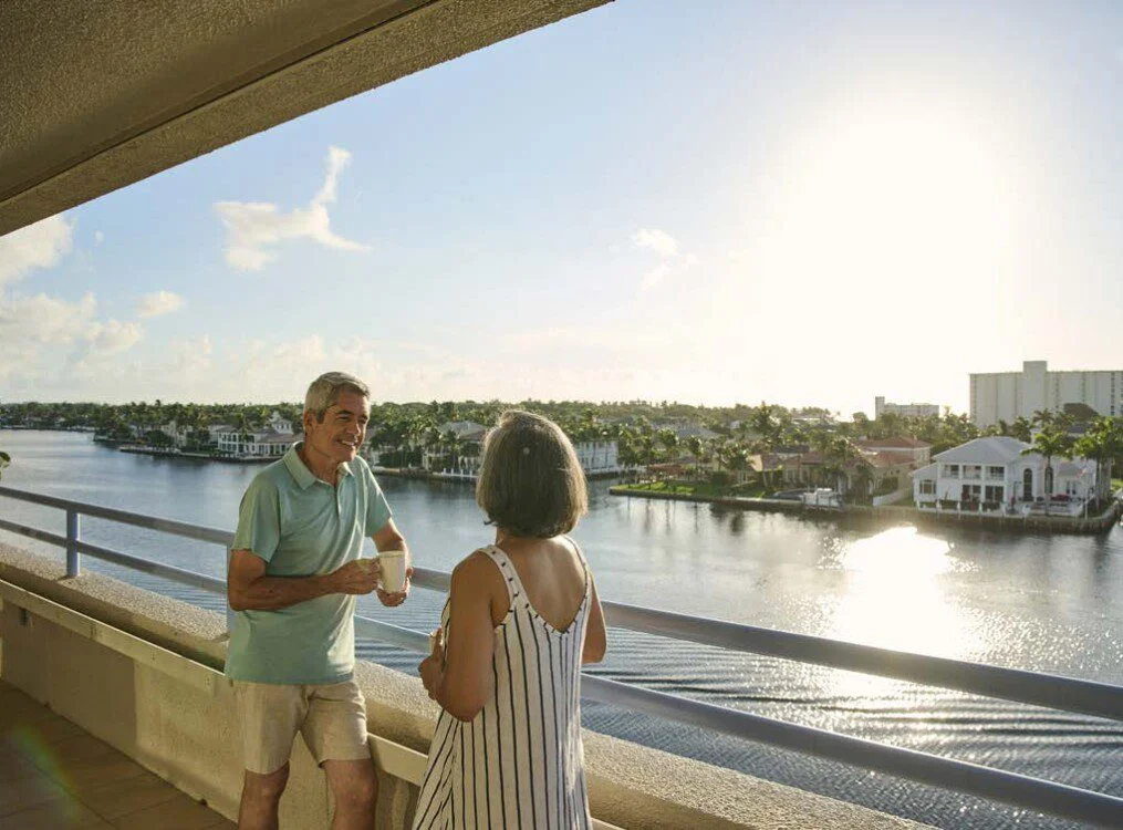 senior couple enjoys coffee while standing by the railing of the balcony Harbour's Edge Senior Living Community, backdropped by water