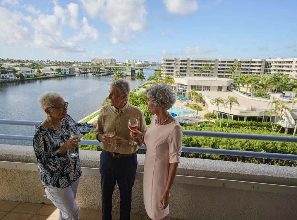 three senior friends enjoy drinks and converse on the balcony overlooking Harbour's Edge Senior Living Community and its namesake harbor