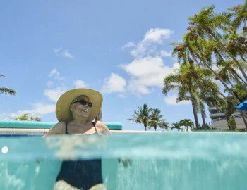 half-underwater shot of smiling senior woman in wide-brimmed hat and sunglasses leaning against the edge of an outdoor resort pool