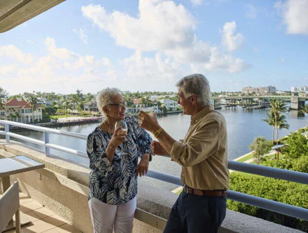 senior man and woman enjoy drinks while standing outside near the railing of the balcony at Harbour's Edge, backdropped by beautiful scenery