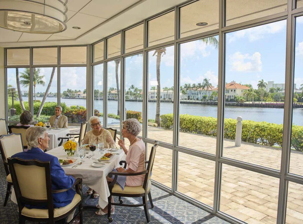 two tables of seniors converse while enjoying dinner and wine inside, backdropped by glass walls and the waterfront views at Harbour's Edge