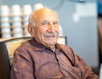 close-up of smiling senior man with mustache looking directly at the camera while seated at a dining table
