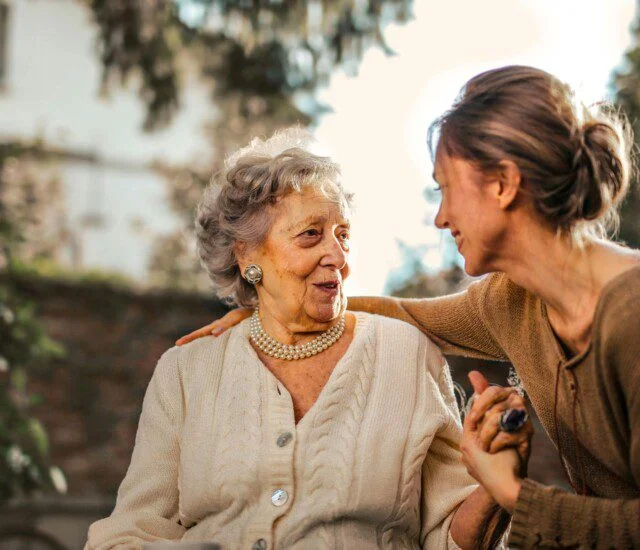 A woman embraces an elderly lady outdoors with trees and sunlight behind them.