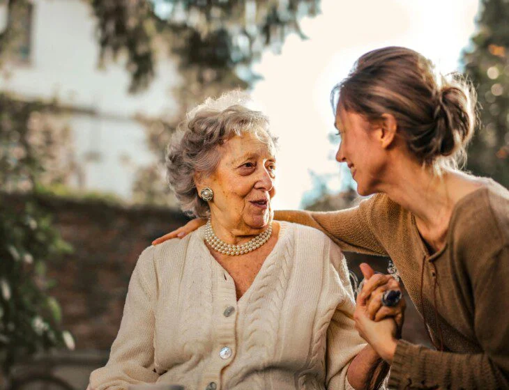 A woman embraces an elderly lady outdoors with trees and sunlight behind them.