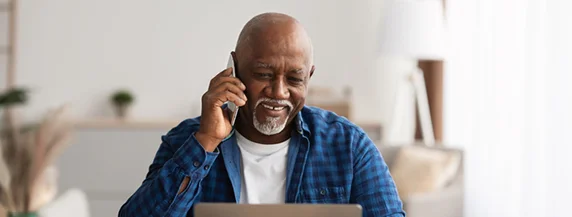 Mature African American Freelancer Man Talking On Cellphone And Using Laptop Computer Sitting At Desk In Modern Office. Senior Businessman Chatting Via Mobile Phone. Business Communication Mature African Man Talking On Cellphone Using Laptop In Office