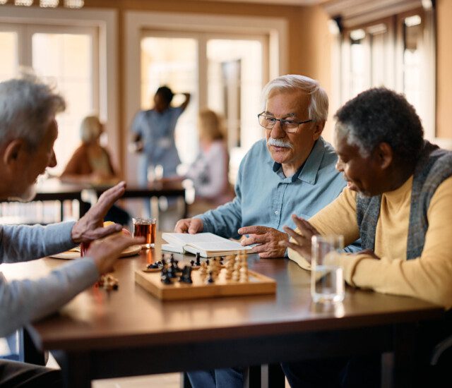 Mature man reading book and communicating with male friends while they are playing chess at retirement community.