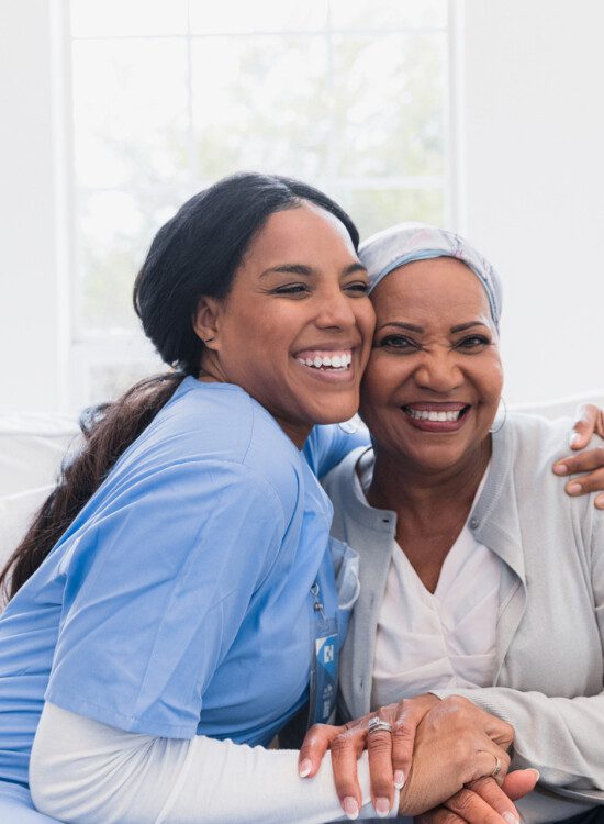 senior woman smiles and hugs her caregiver while seated on a couch in her apartment