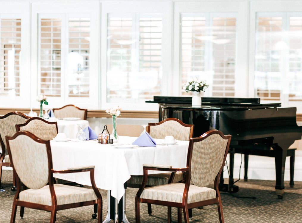 dining tables draped with crisp white cloths in the dining room at Harbour's Edge Senior Living Community, a grand piano in the background