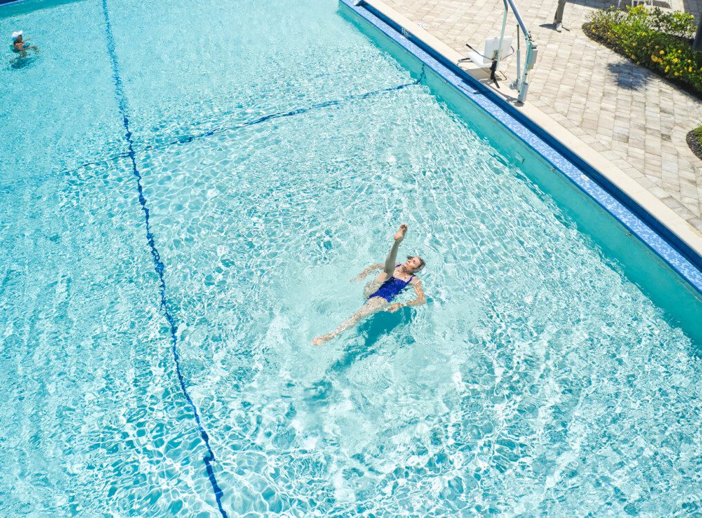 aerial view of senior woman kicking her leg up while swimming on her back in an outdoor, resort-style pool at her senior living community