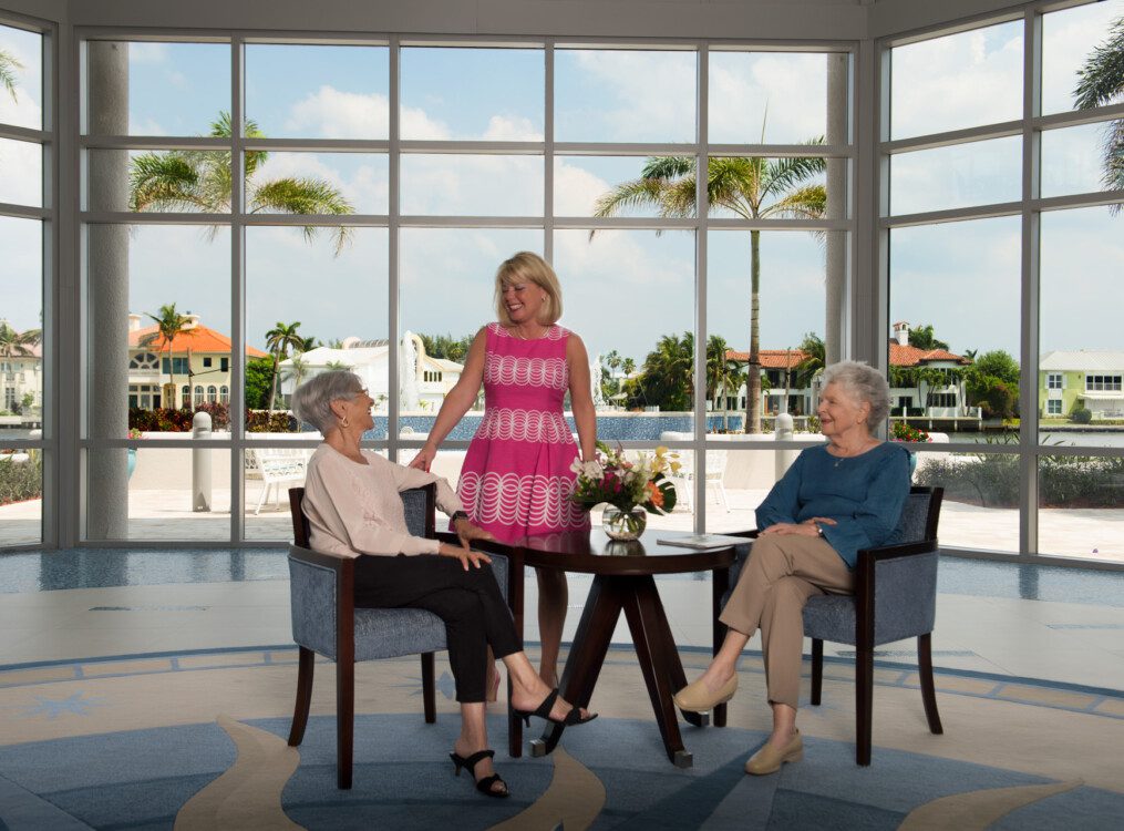 two senior women seated at a small table in front of glass walls speaking with a woman in a pink dress, backdropped by the community pool