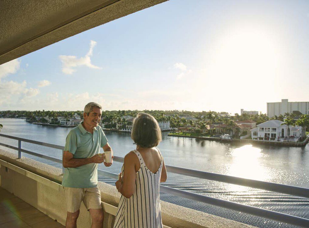smiling senior couple enjoys coffee on the balcony, looking out over the water and landscape of Harbour's Edge Senior Living Community