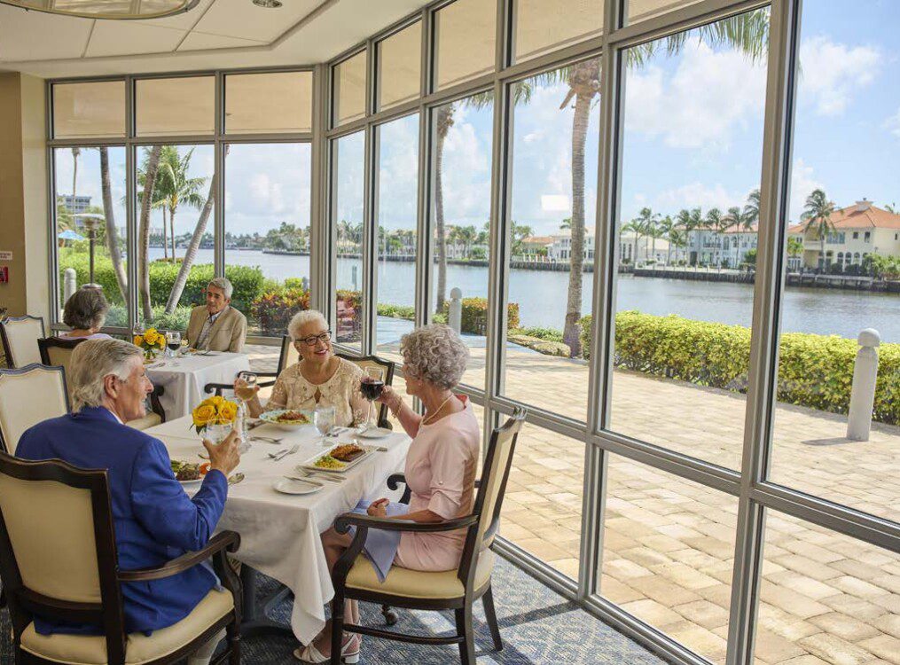 two tables of seniors converse while enjoying dinner and wine inside, backdropped by glass walls and the waterfront views at Harbour's Edge