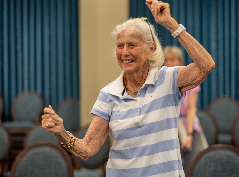 smiling senior woman in striped polo shirt lifts her arms and dances during a fitness class at her senior living community
