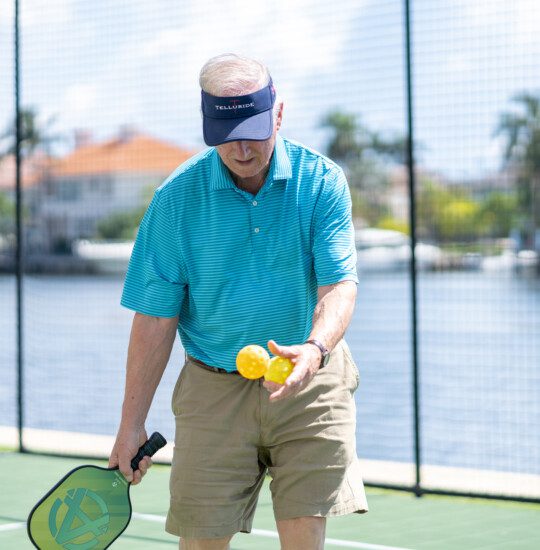 senior man wearing visor hat prepares to serve during an outdoor, waterfront game of pickleball at his senior living community