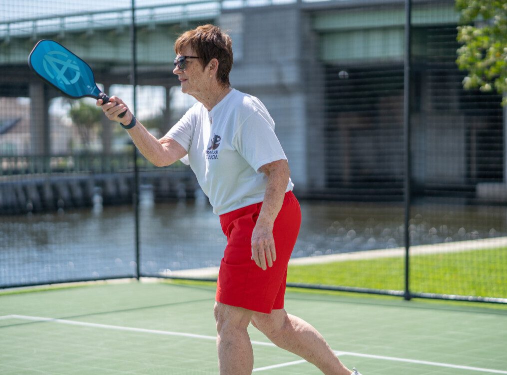 senior woman in sunglasses swings paddle during an outdoor game of pickleball at her senior living community