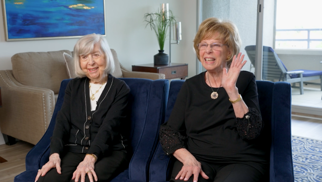 senior woman and her adult daughter smile and sit on black chairs side-by-side for an interview