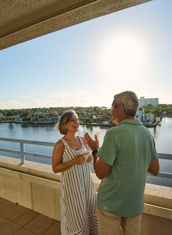 smiling senior couple enjoys coffee on the balcony, looking out over the water and landscape of Harbour's Edge Senior Living Community