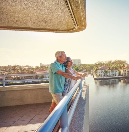 smiling senior couple leans against balcony railing, looking out over the water and landscape of Harbour's Edge Senior Living Community