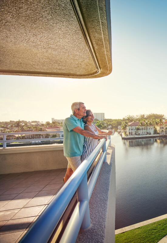 smiling senior couple leans against balcony railing, looking out over the water and landscape of Harbour's Edge Senior Living Community