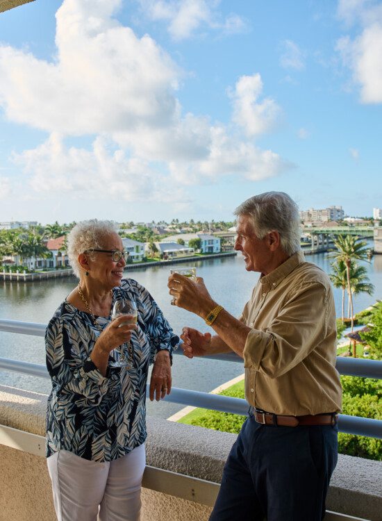 senior man and woman smile while enjoying drinks on the dining room balcony overlooking Harbour's Edge and its namesake harbor