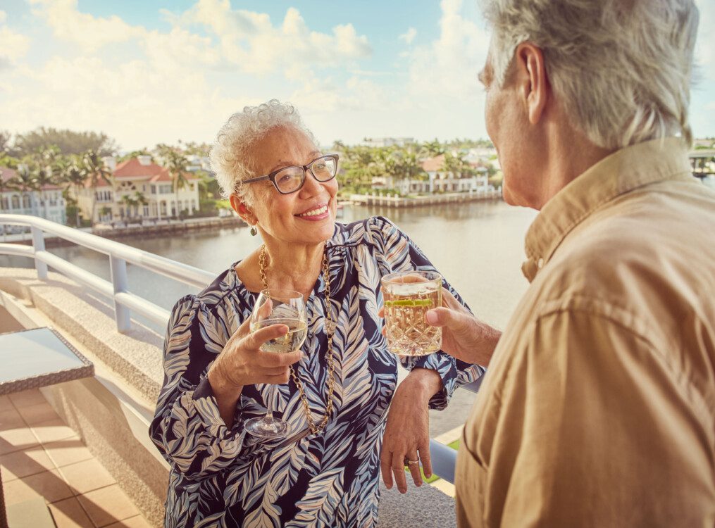 senior man and woman smile while enjoying drinks on the dining room balcony overlooking Harbour's Edge and its namesake harbor