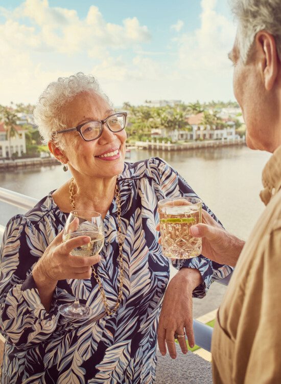 senior man and woman smile while enjoying drinks on the dining room balcony overlooking Harbour's Edge and its namesake harbor