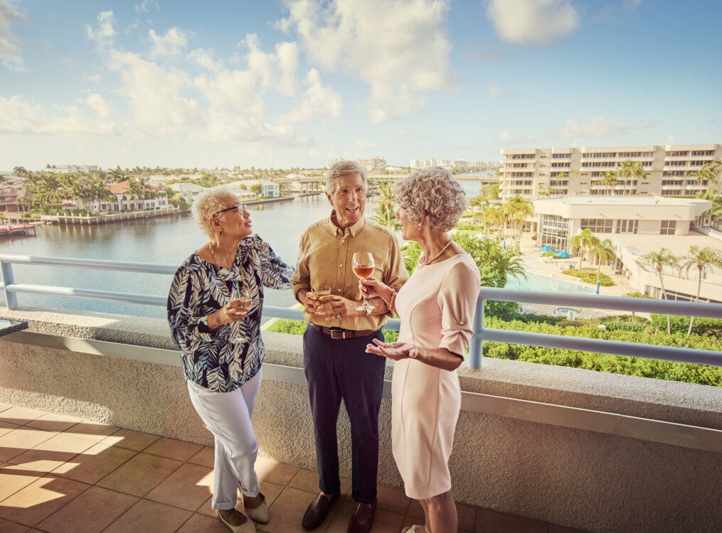 three senior friends enjoy drinks and converse on the balcony overlooking Harbour's Edge Senior Living Community and its namesake harbor