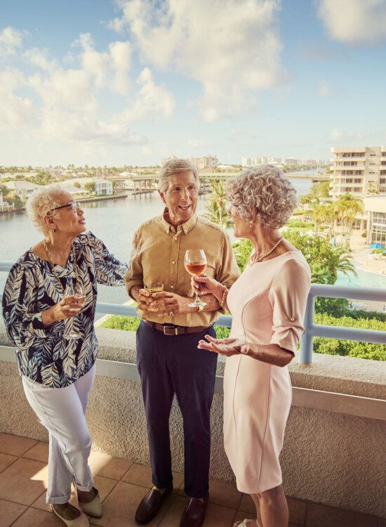 three senior friends enjoy drinks and converse on the balcony overlooking Harbour's Edge Senior Living Community and its namesake harbor