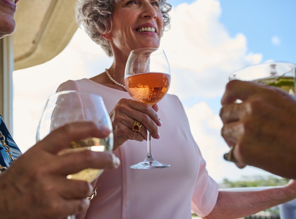 close-up of three senior friends smiling and holding stemmed wine glasses, backdropped by sunny blue skies
