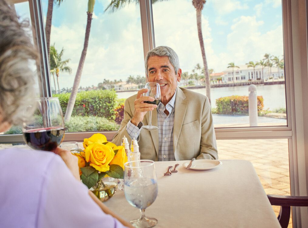 senior man in tan business suit smiles while sipping wine at an upscale dinner with his wife at Harbour's Edge Senior Living Community