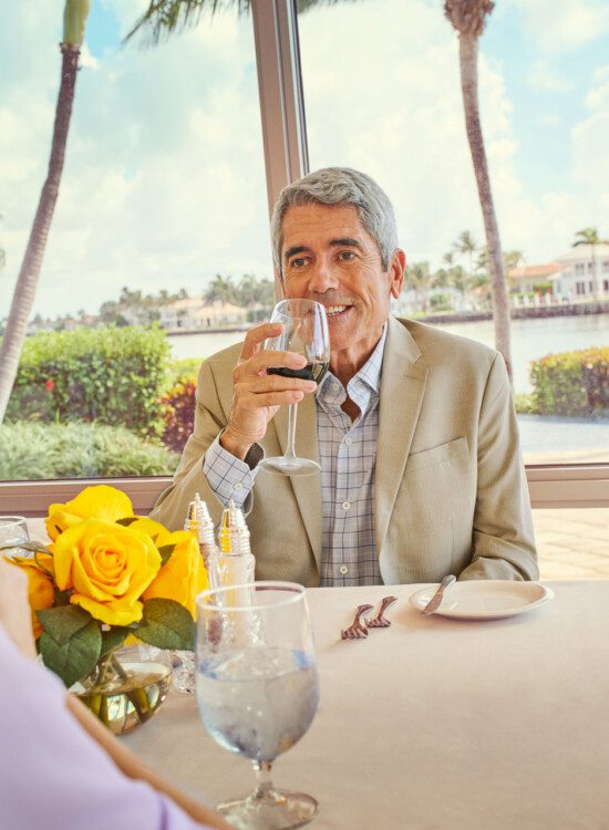 senior man in tan business suit smiles while sipping wine at an upscale dinner with his wife at Harbour's Edge Senior Living Community