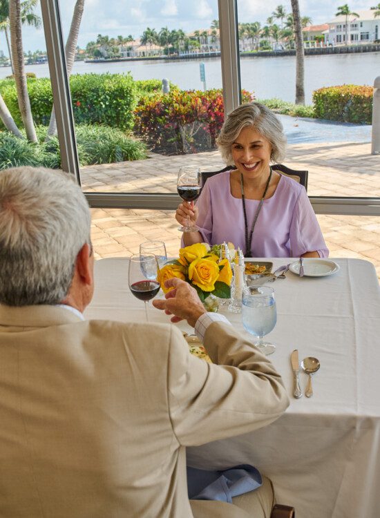 smiling senior couple raise wine glasses for a toast while dining in the upscale venue at Harbour's Edge, backdropped by scenic water views