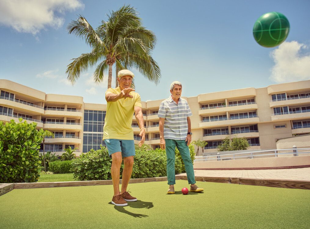 two smiling senior men in casual clothes play bocce ball outdoors at Harbour's Edge Senior Living Community in Delray Beach, FL