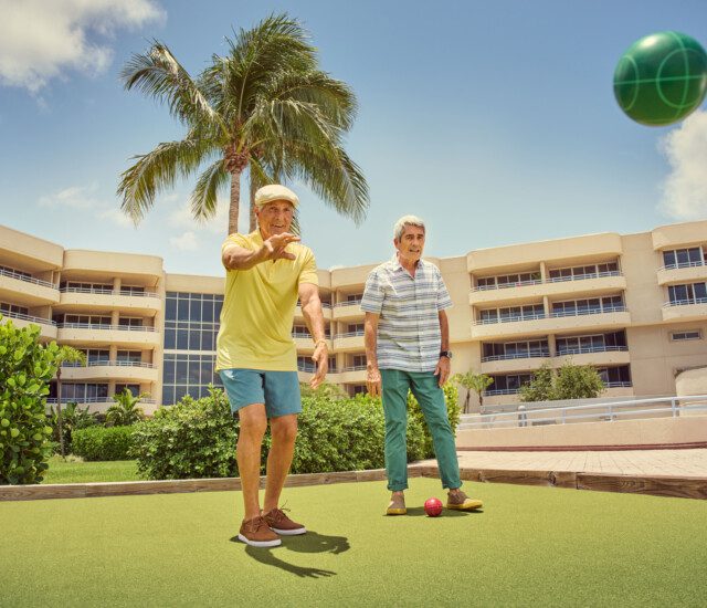 two smiling senior men in casual clothes play bocce ball outdoors at Harbour's Edge Senior Living Community in Delray Beach, FL