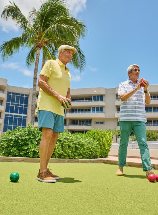 two smiling senior men in casual clothes play bocce ball outdoors at Harbour's Edge Senior Living Community in Delray Beach, FL
