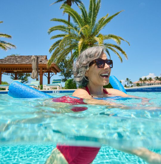 smiling senior woman in sunglasses uses a pool noodle to float in the outdoor pool at Harbour's Edge, backdropped by palm trees