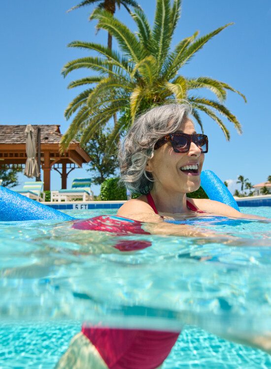 smiling senior woman in sunglasses uses a pool noodle to float in the outdoor pool at Harbour's Edge, backdropped by palm trees