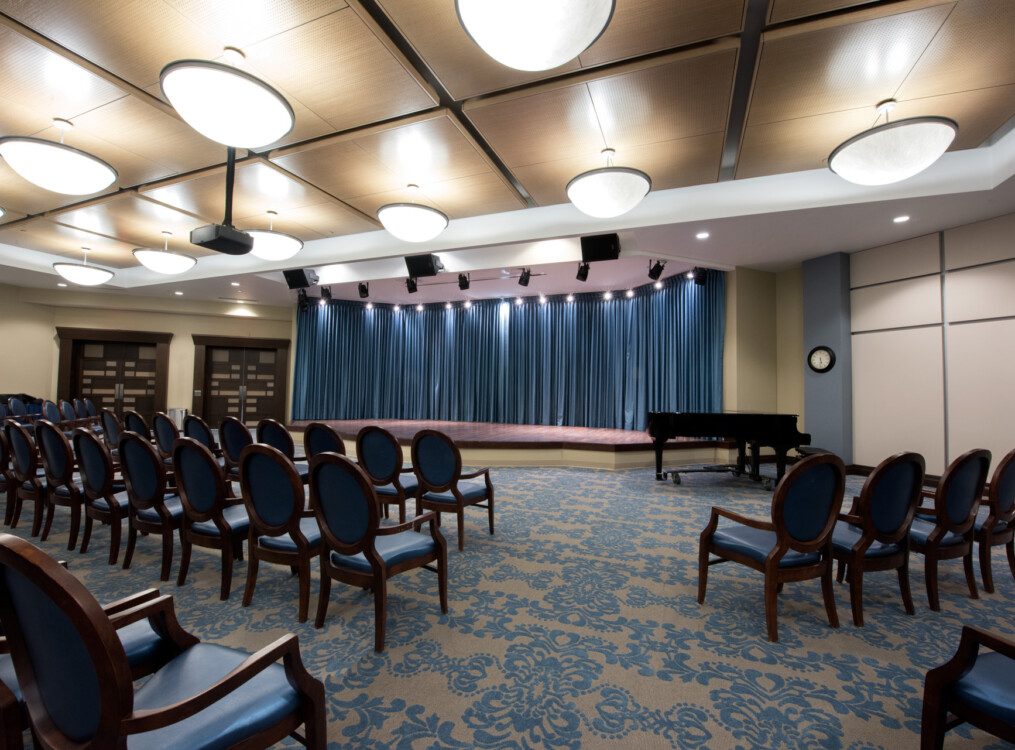 empty theater room with small stage, lights, piano, and rows of chairs at Harbour's Edge Senior Living Community