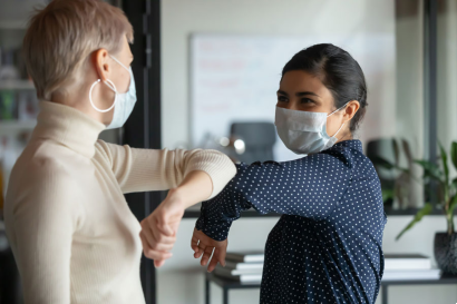 two women greeting each other by touching elbows while wearing face masks