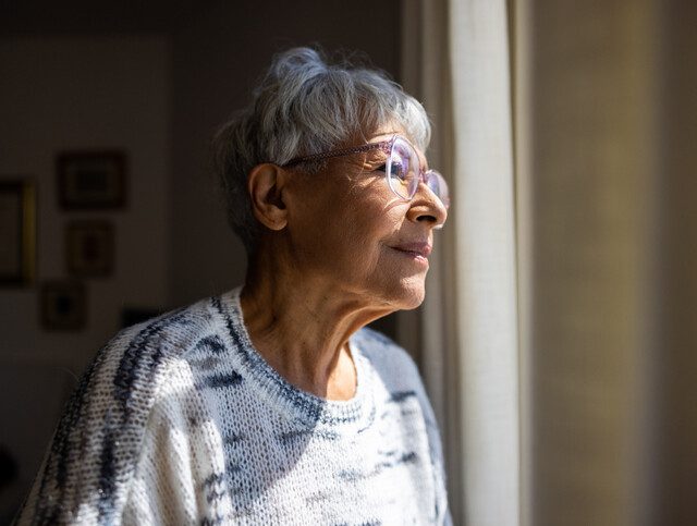 Older adult woman thoughtfully looks outside her window.
