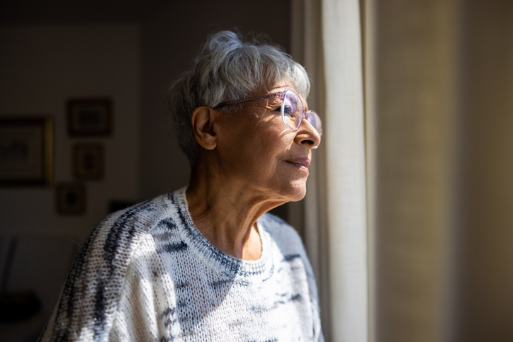 Older adult woman thoughtfully looks outside her window.