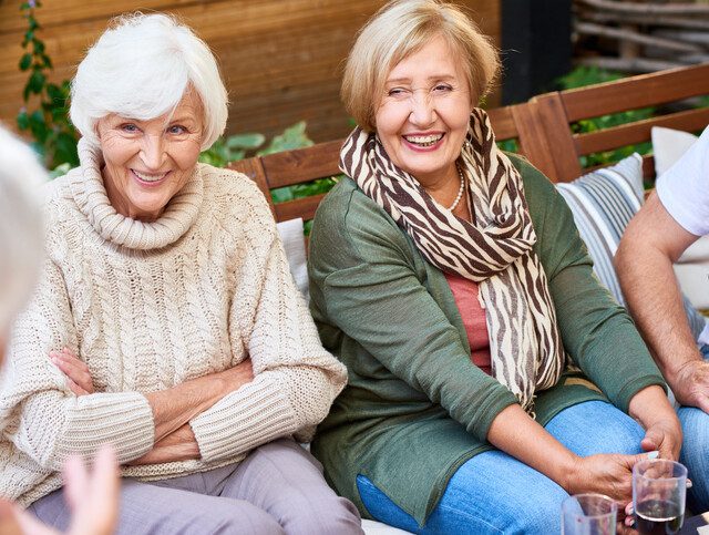 Two older adult women seated and chatting with friends outside.