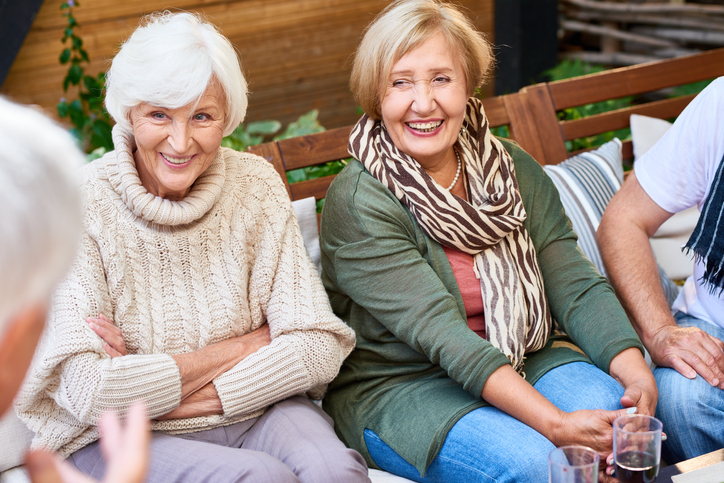 Two older adult women seated and chatting with friends outside.