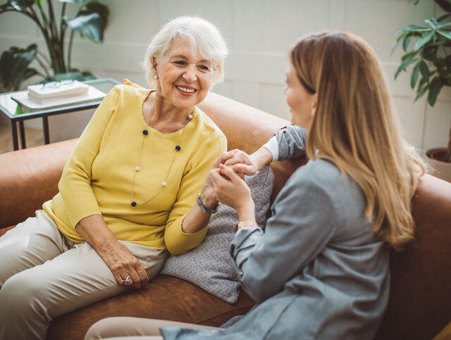 Older adult woman engages in conversation with adult daughter on couch.